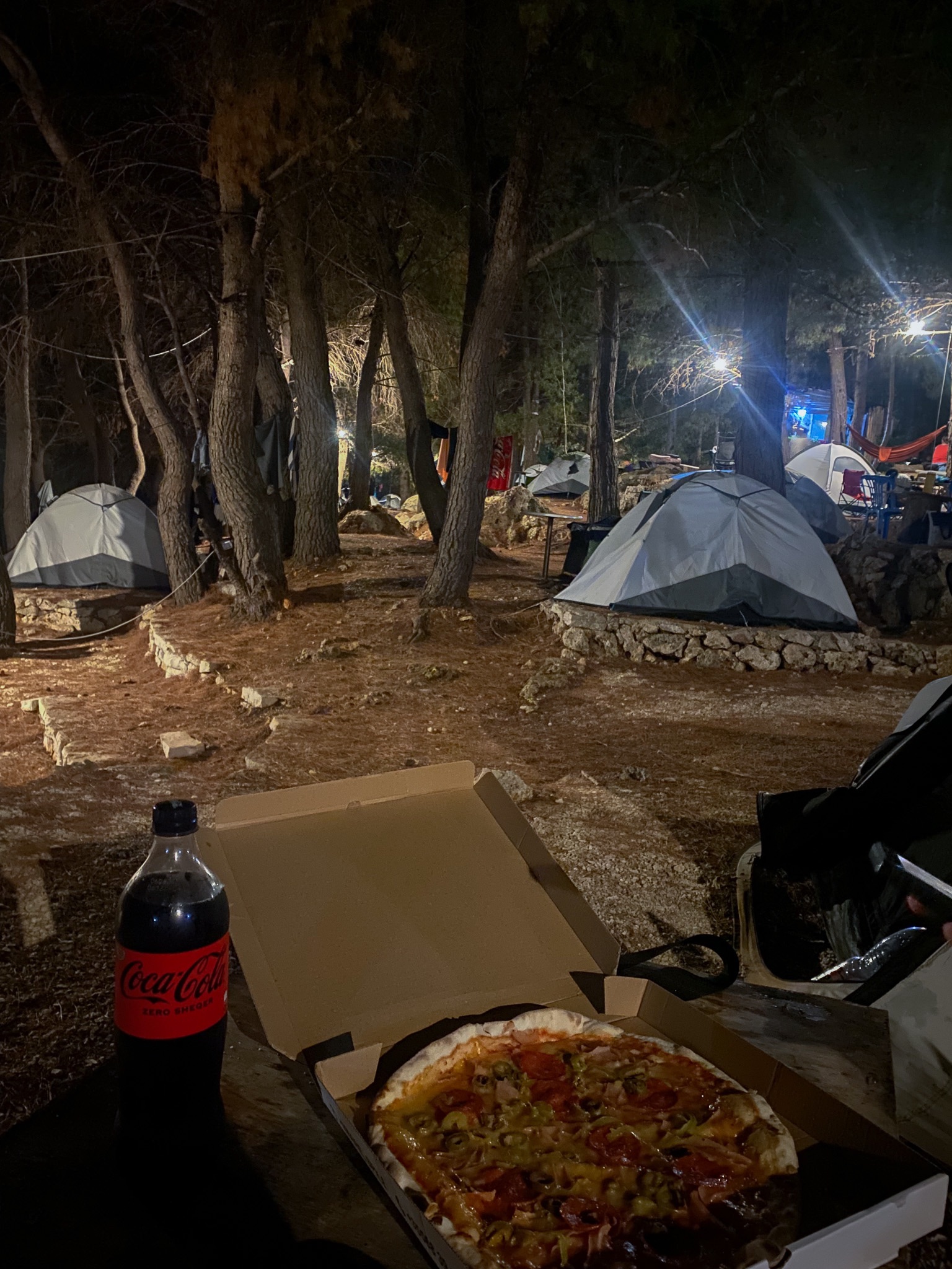 A takeout pizza box and a bottle of Coca-Cola on a wooden table at night, with illuminated camping tents nestled under pine trees at Pine Side Camp in Himarë.