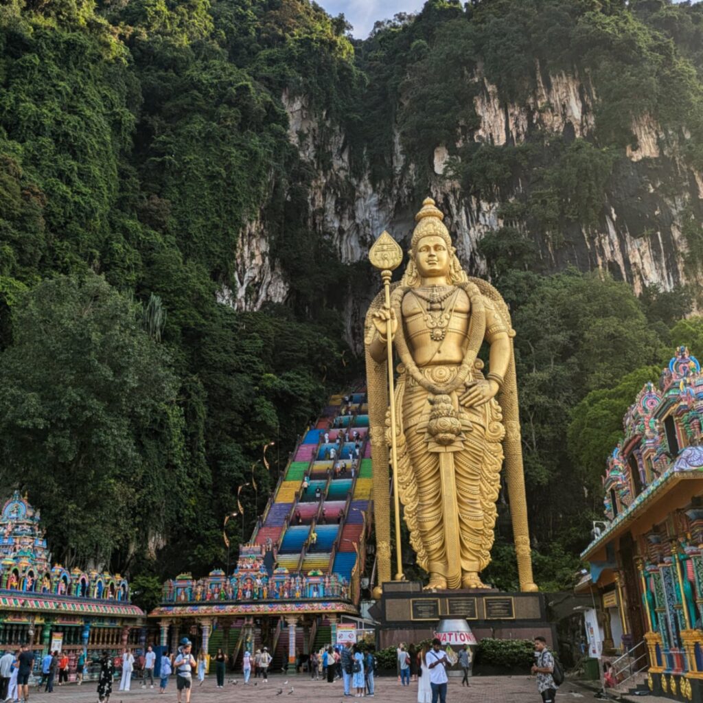 The budget-friendly Batu Caves entrance in Malaysia, featuring the towering golden statue of Lord Murugan standing prominently on the right. To the left, a steep, vibrant rainbow-colored staircase ascends into the massive limestone cave opening, surrounded by lush green cliffs and tourists gathered at the base.