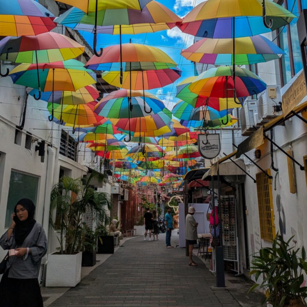 A narrow pedestrian alleyway shaded by a vibrant canopy of suspended, multi-colored rainbow umbrellas overhead. Pedestrians walk along the path in Georgetown, Penang, Malaysia.