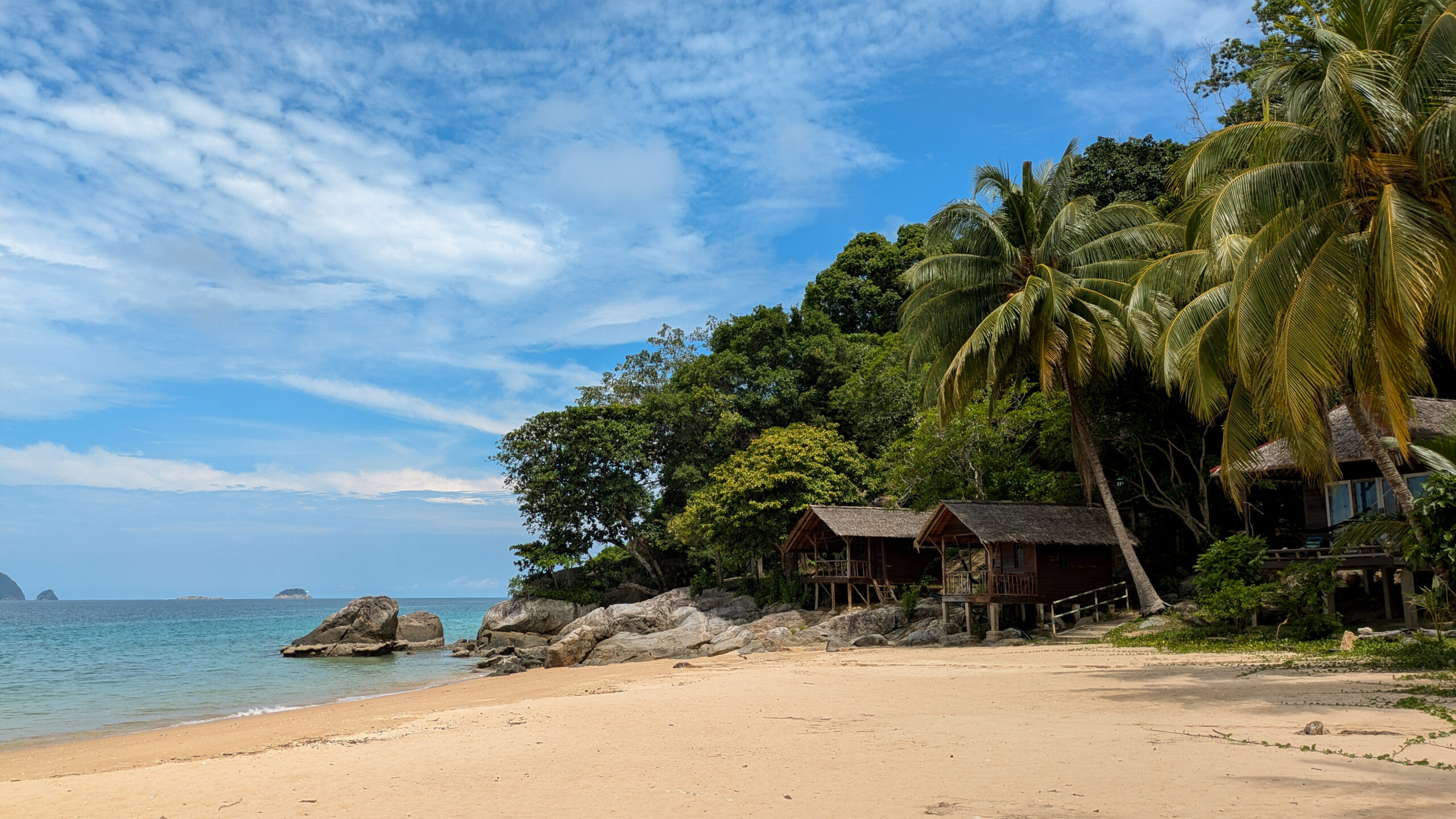A serene tropical beach on the Perhentian Islands, Malaysia featuring a wide stretch of golden sand, calm turquoise ocean water, and several small wooden chalets built on stilts along the edge of a lush, forested hill. Large granite rocks sit near the shoreline, and tall coconut palm trees frame the right side of the view under a bright blue sky with scattered white clouds.