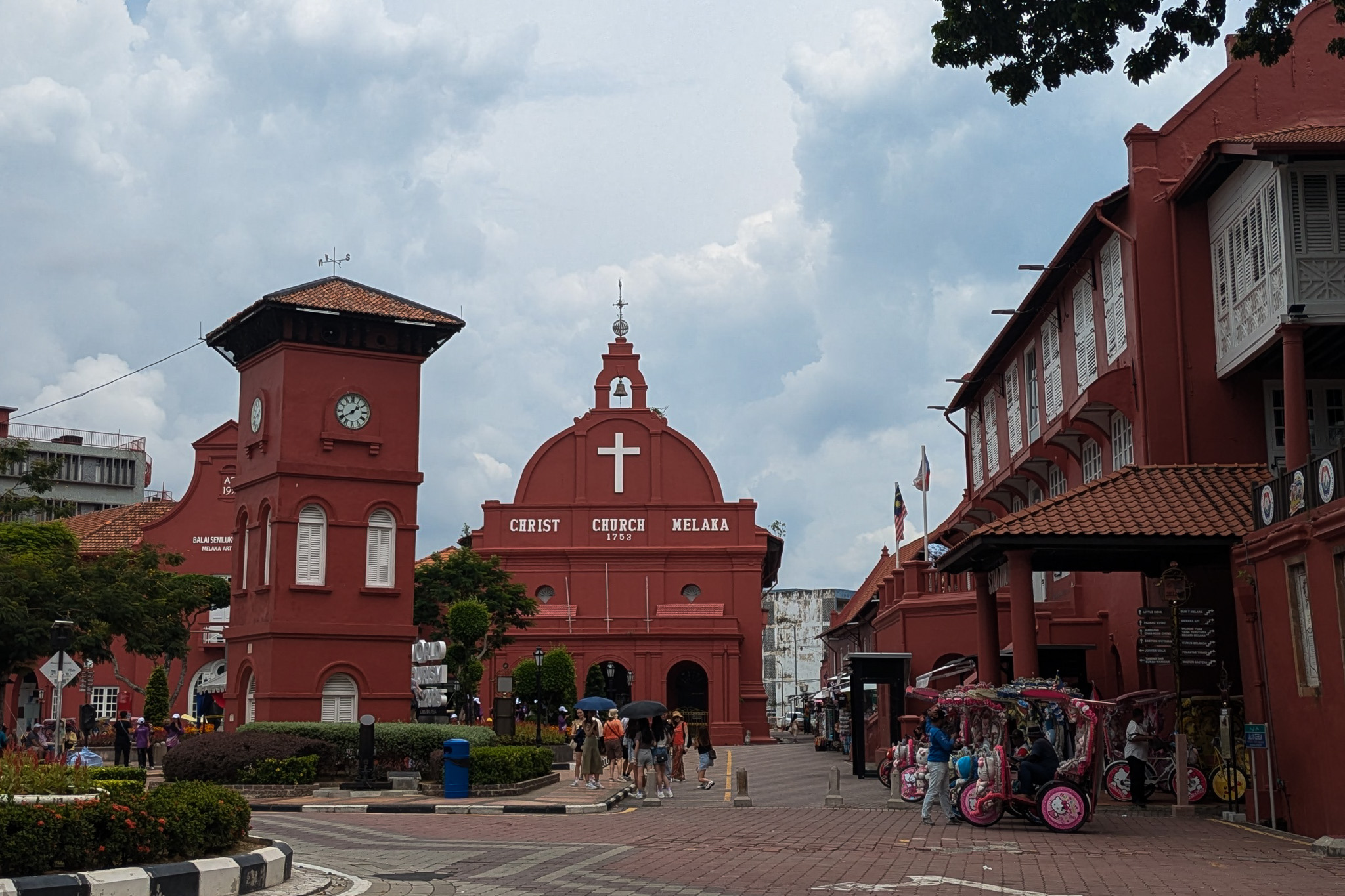 The historic terracotta-red Christ Church and neighboring clock tower stand in a paved square with tourists and decorated trishaws under a cloudy sky in Malacca, Malaysia