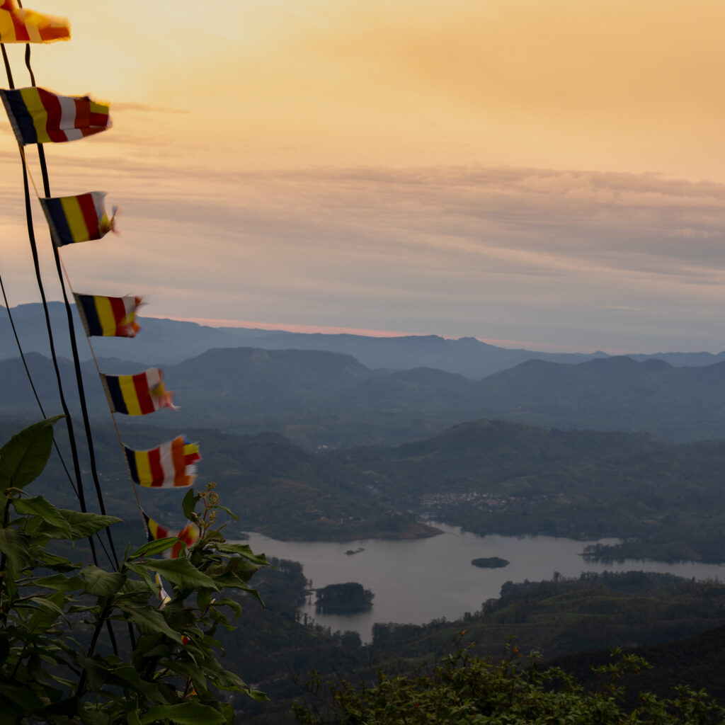 Sunrise view from Adam's Peak (Sri Pada) Sri Lanka with Buddhist prayer flags over misty mountain valleys and reservoir.