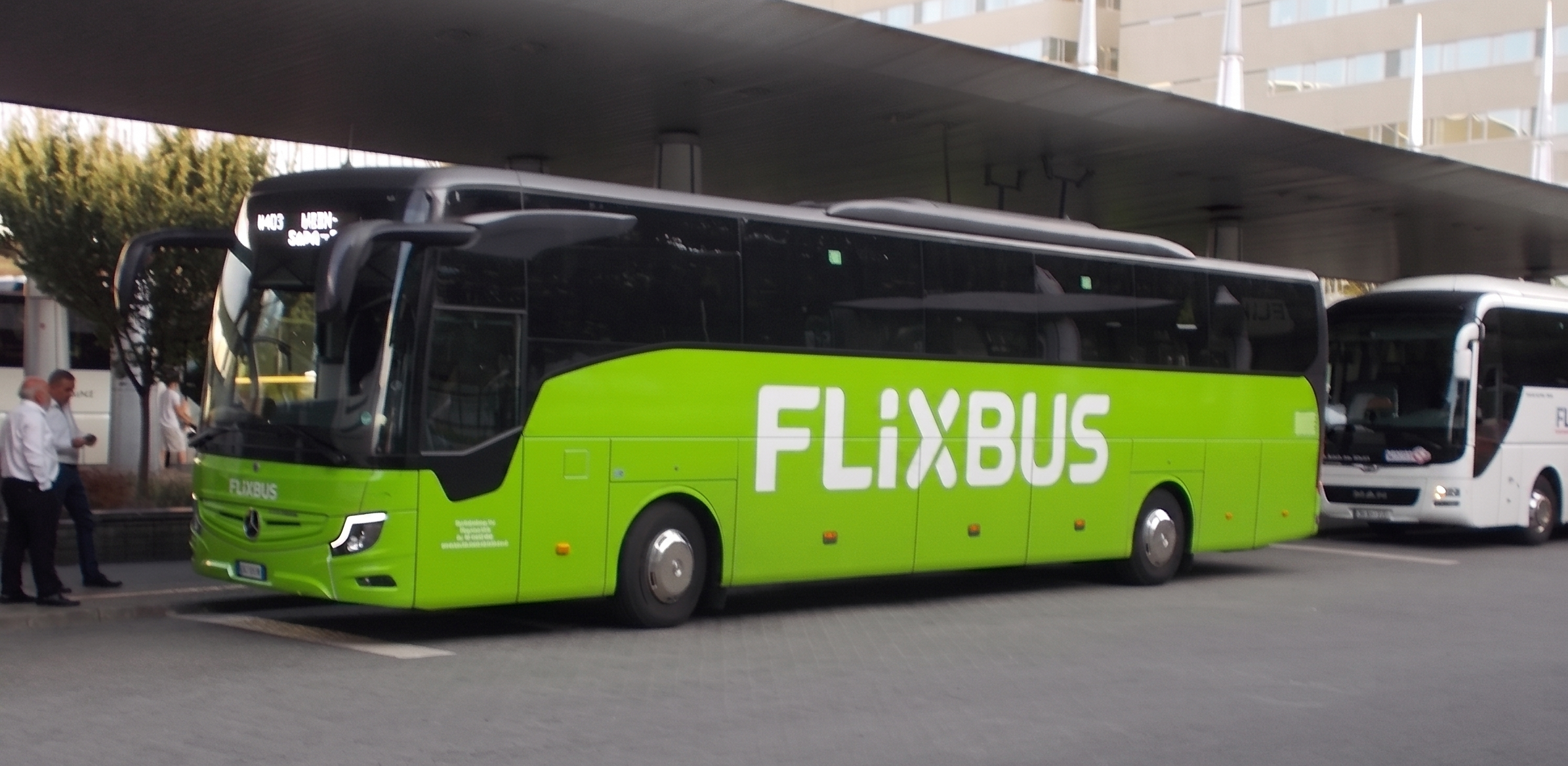 A photo of a bright green flixbus parked at a European bus station with 2 bus workers at the front and a white bus parked behind it