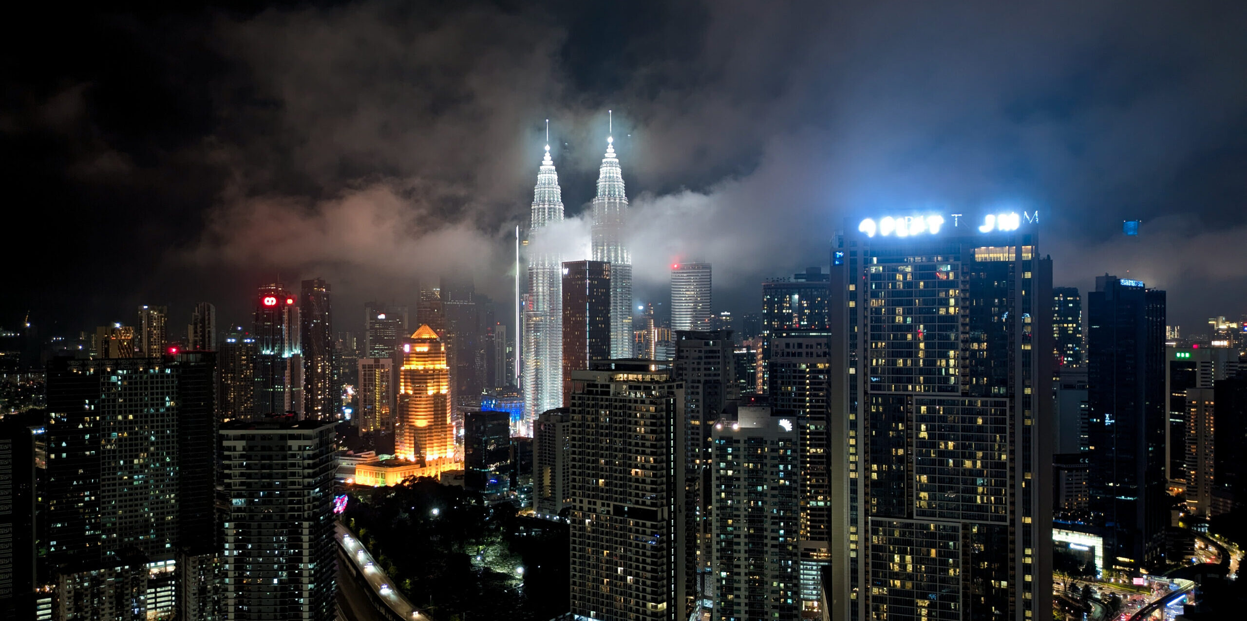 The Kuala Lumpur skyline at night with the Petronas Towers lit bright and misty clouds floating through the skyscrapers