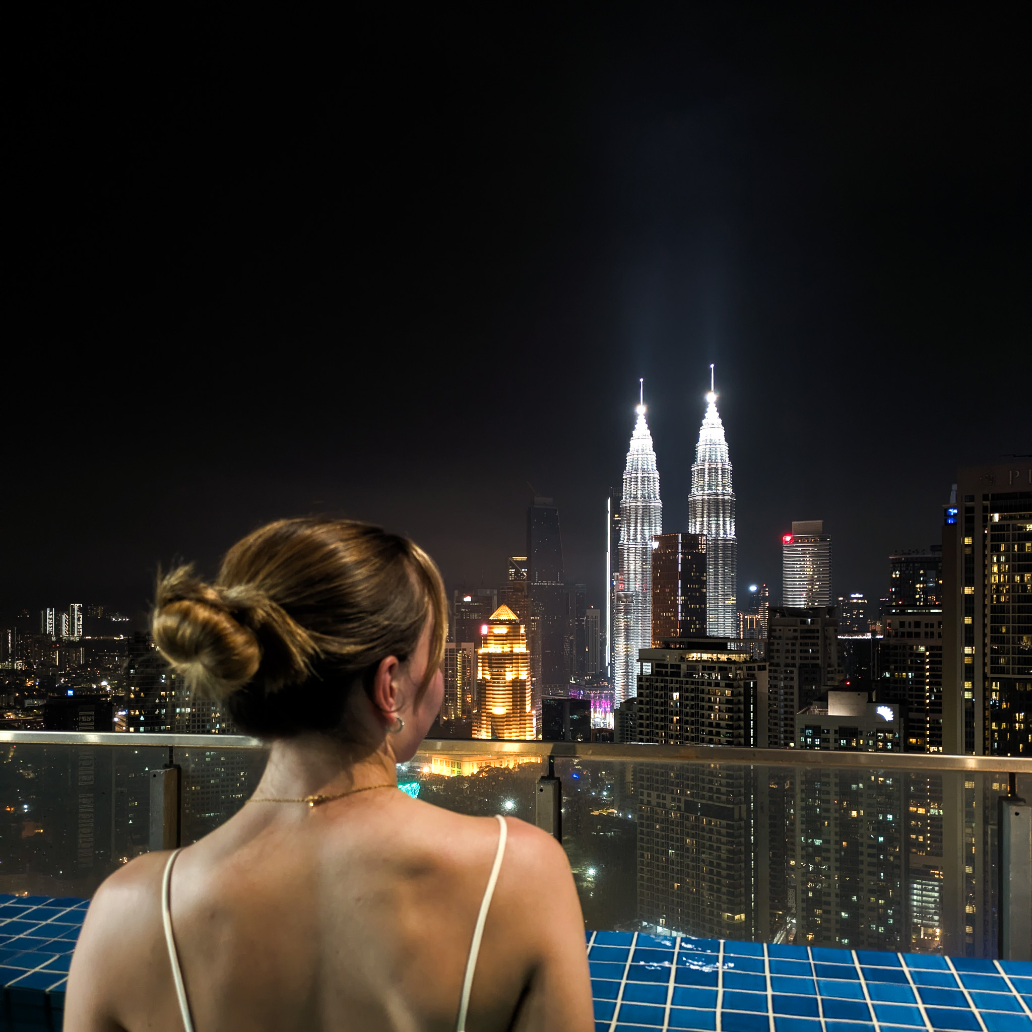 A woman in a swimming pool looking out over the night sky of Kuala Lumpur with the Petronas Twin Towers glowing in the distance