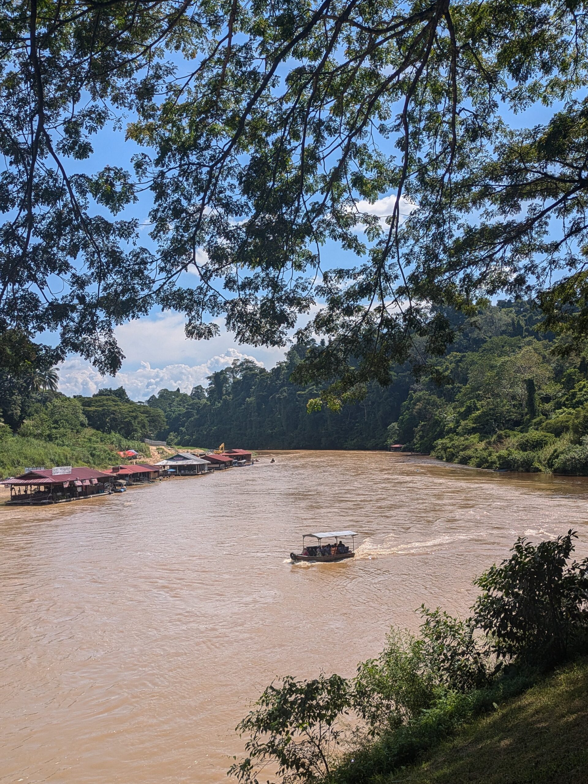 A small boat navigating a wide brown jungle river past floating wooden docks in Taman Negara, Malaysia.
