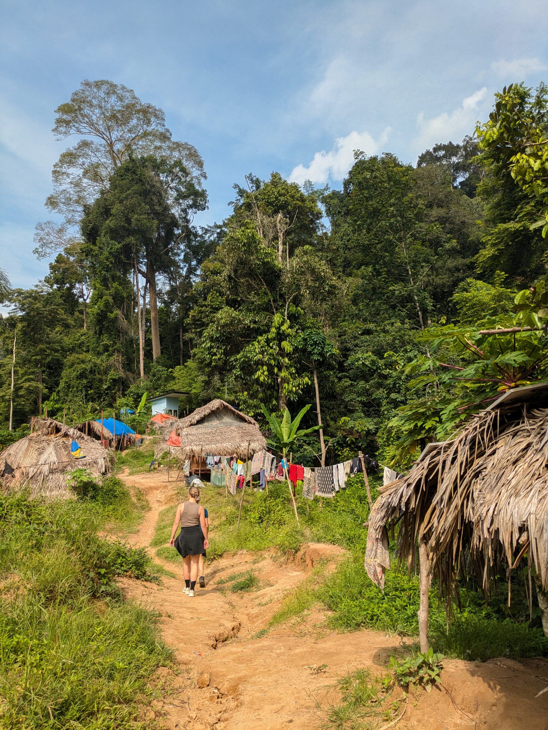 A woman walking along a dirt path past thatched-roof huts in a rural jungle village in Taman Negara, Malaysia.