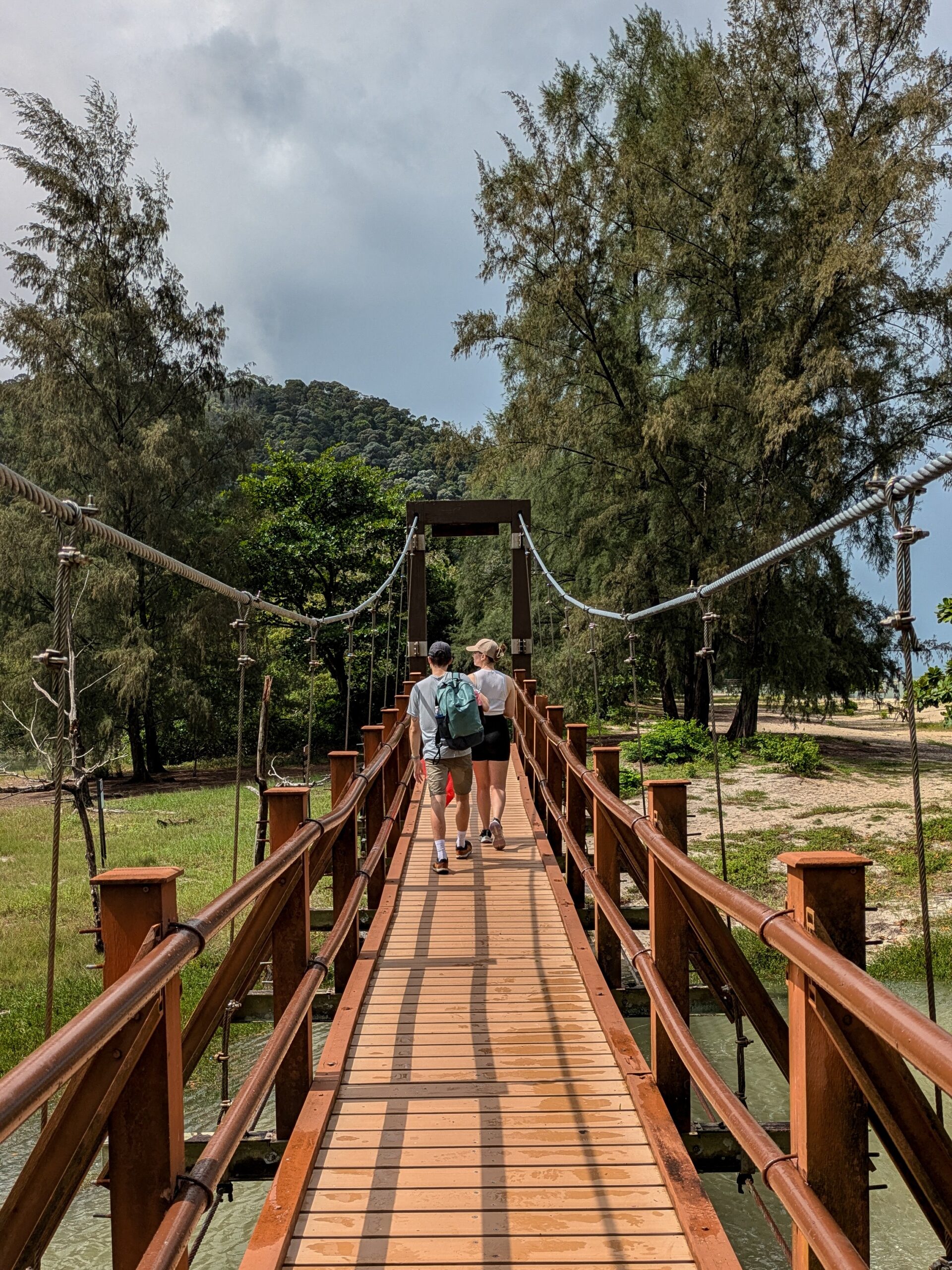 Two hikers walking away across a wooden suspension bridge toward a sandy path lined with tall trees and a forested hill in Penang National Park, Malaysia.