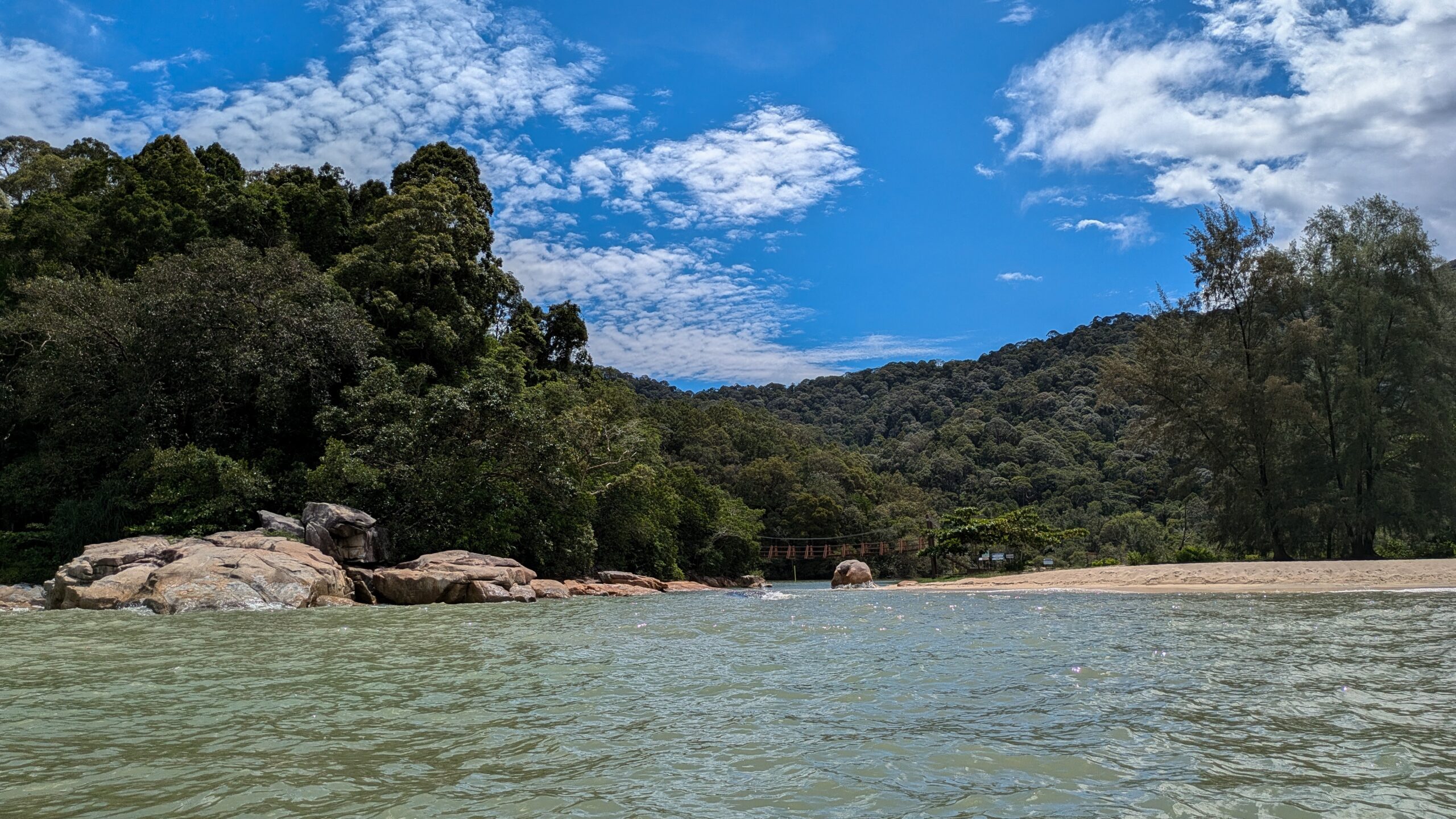 Coastal view of a sandy beach and a small suspension bridge nestled in thick green woods of Penang National Park, Malaysia.