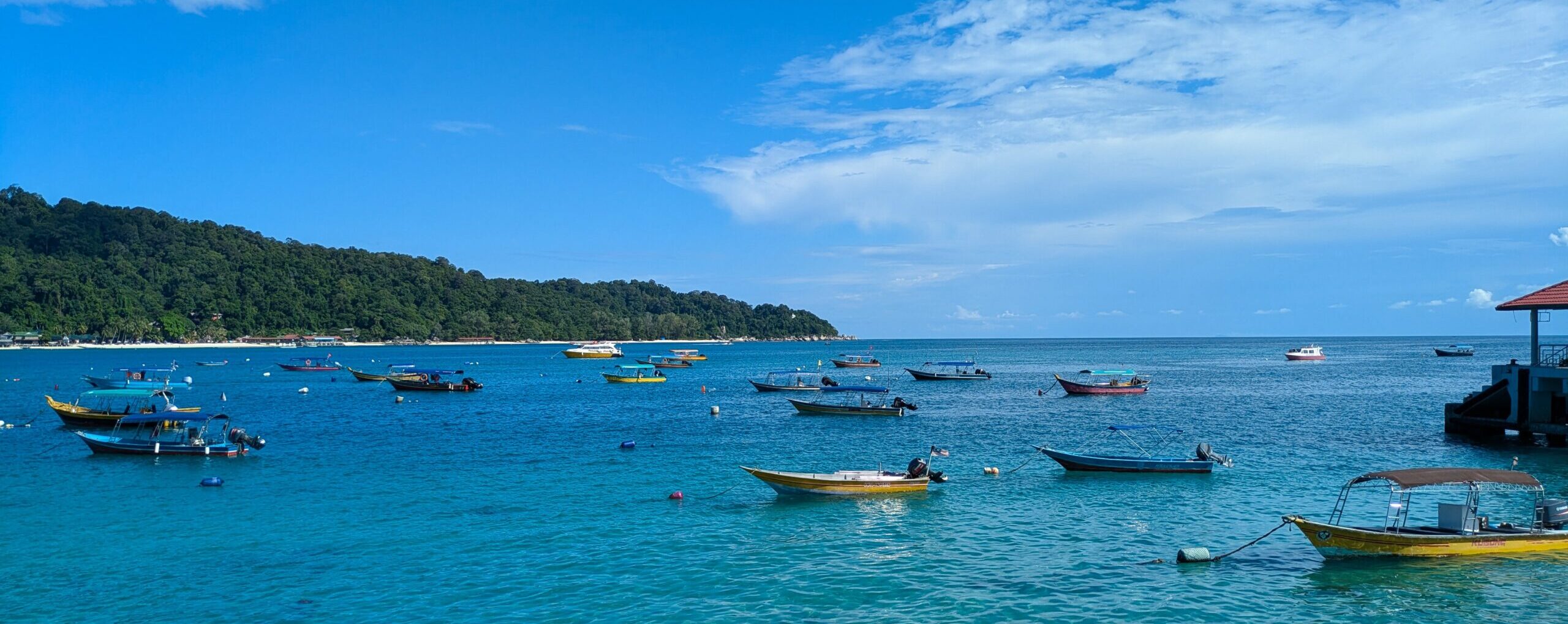 Multiple small boats anchored in clear turquoise water off the coast of a lush, jungle-covered tropical island of the Perhentian Islands, Malaysia