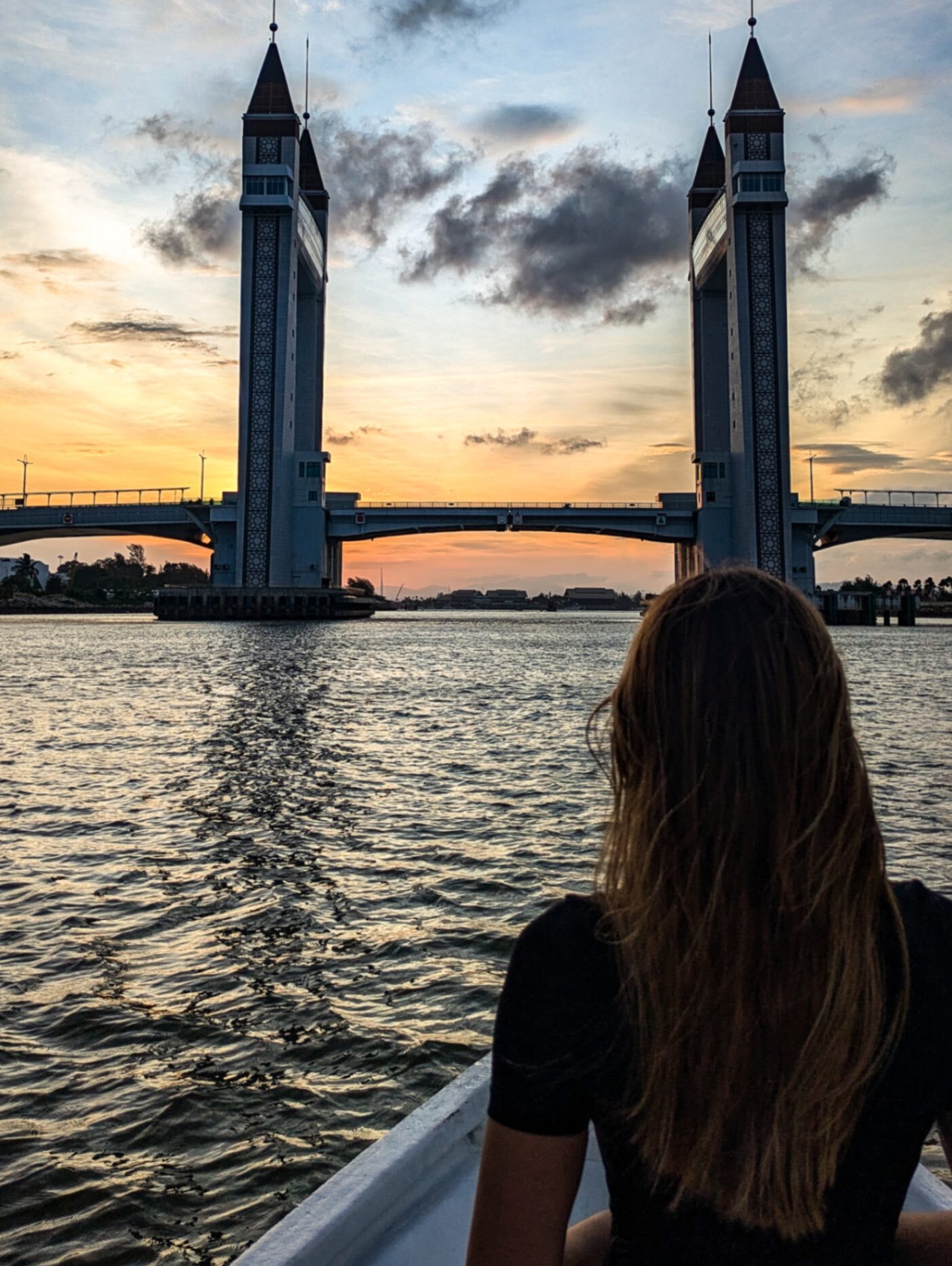 A women on a boat on water with an orange sunset sky beyond a drawbridge in Kuala Terrenganu, Malaysia