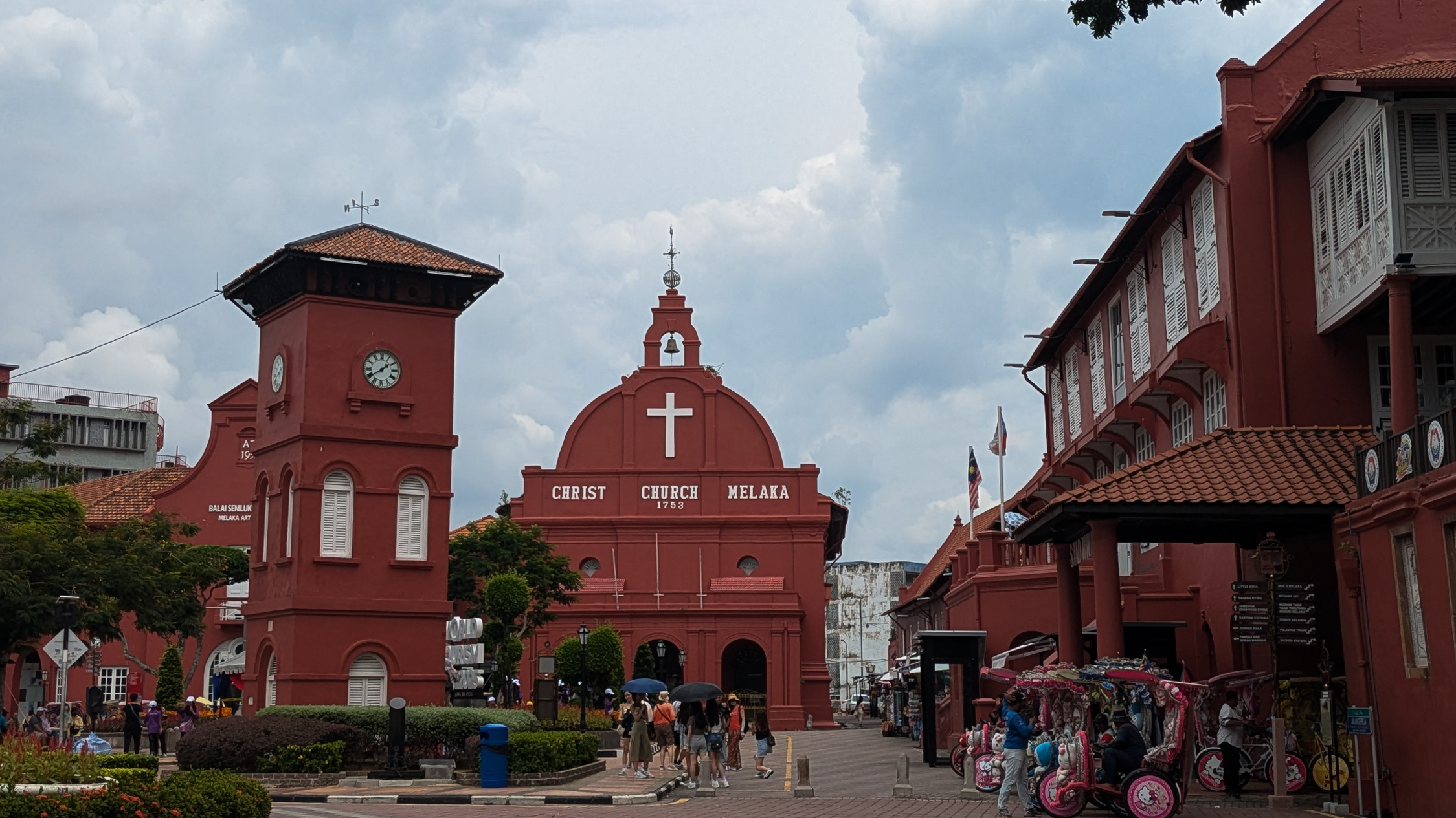 The historic terracotta-red Christ Church and neighboring clock tower stand in a paved square with tourists and decorated trishaws under a cloudy sky in Malacca, Malaysia