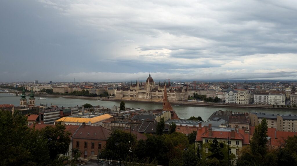 Elevated panoramic view of the Hungarian Parliament Building and the Danube River in Budapest under a dramatic, cloudy sky, captured from Fisherman’s Bastion on the Buda side - lots of great things to do to guide a visitor through a first-time to Budapest
