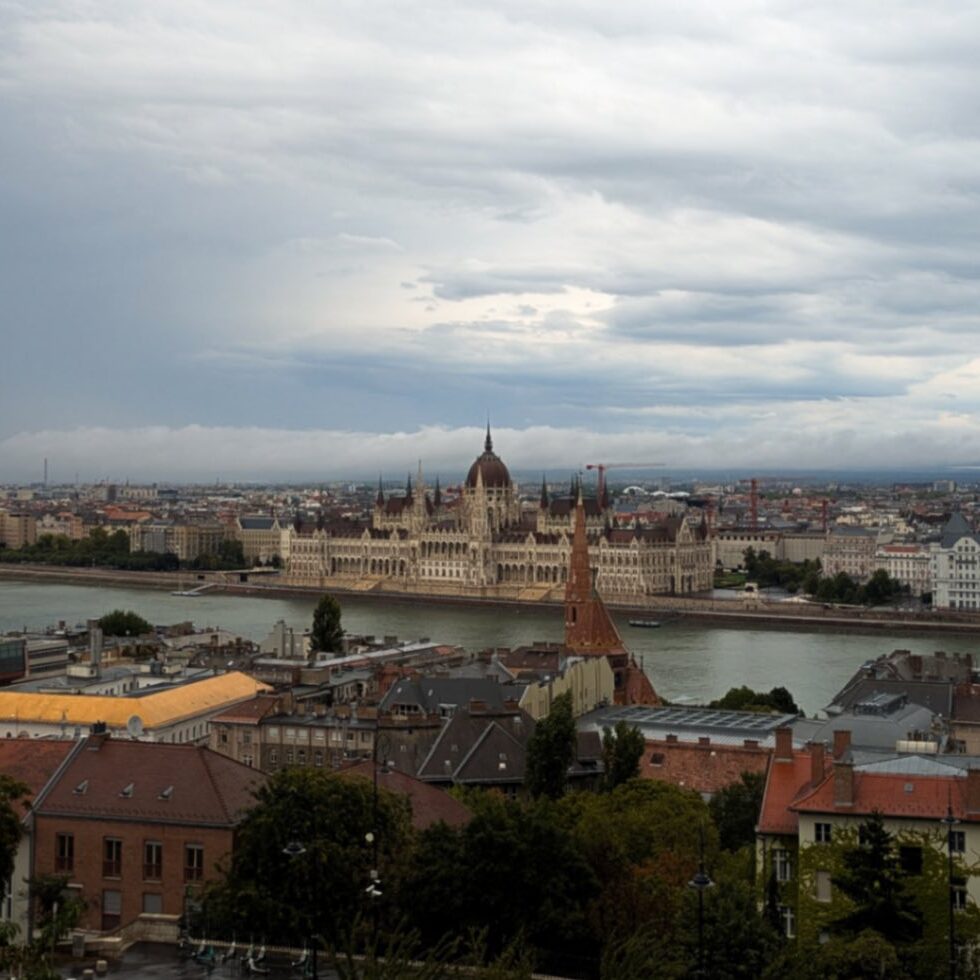 Elevated panoramic view of the Hungarian Parliament Building and the Danube River in Budapest under a dramatic, cloudy sky, captured from Fisherman’s Bastion on the Buda side - lots of great things to do to guide a visitor through a first-time to Budapest