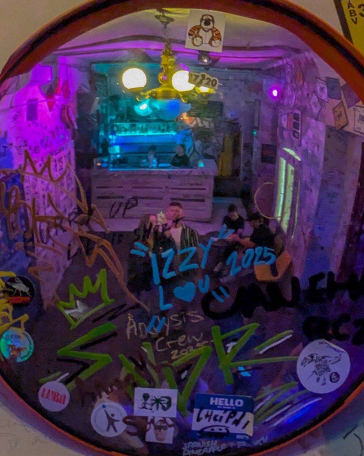 An image of a traffic mirror inside of a bar with two people taking a photo of the stickers and writing on it