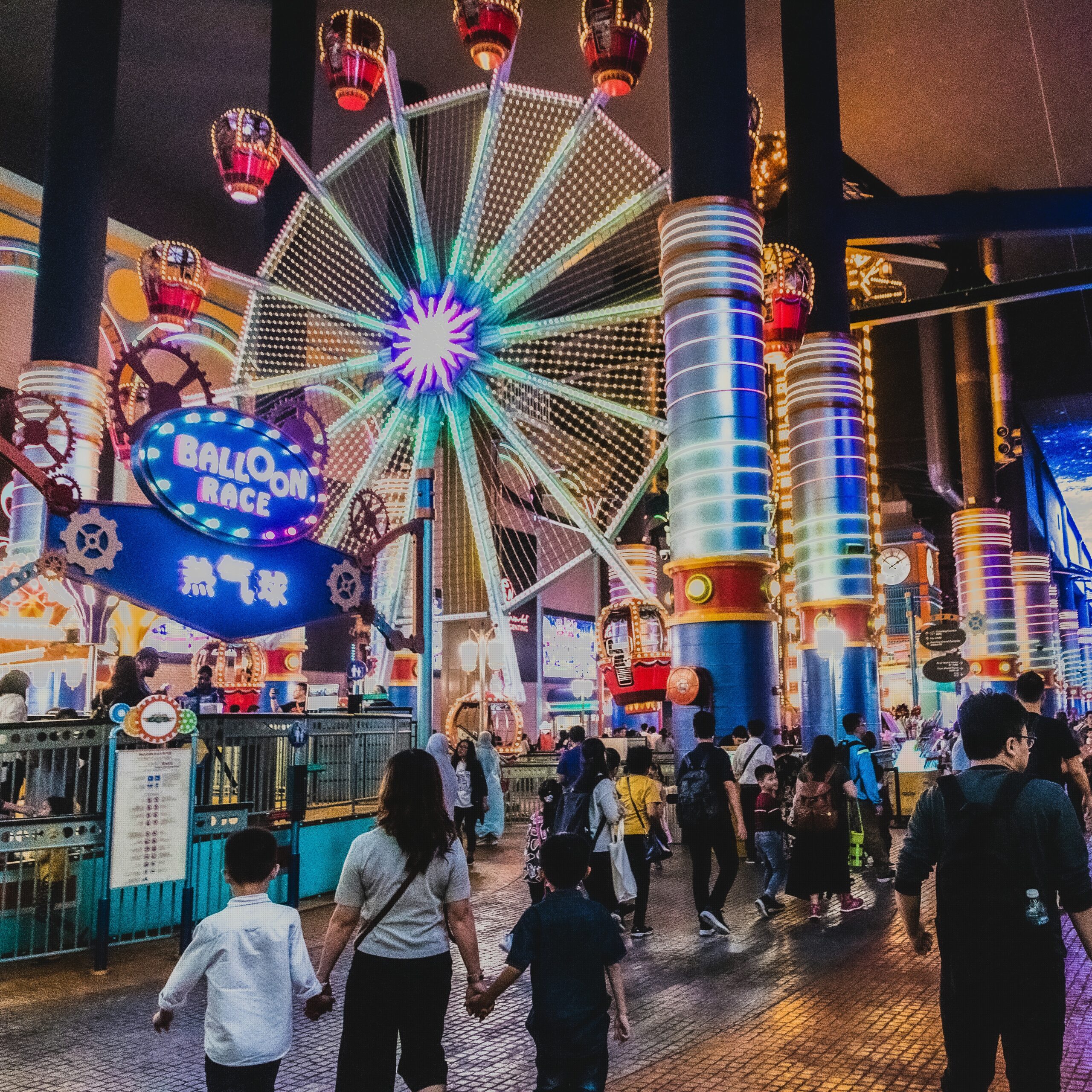 A crowded indoor amusement park at night featuring a large, neon-lit Ferris wheel ride called "Balloon Race" and glowing structural pillars.