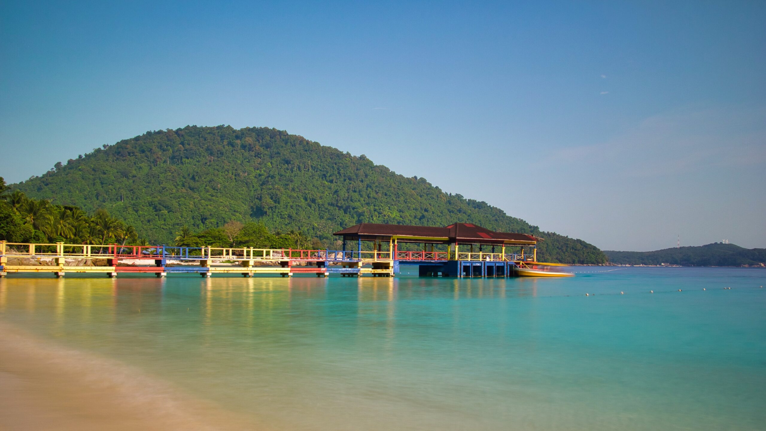 Stunning blue waters under a clear blue sky with a colourful jetty in front of a lush green mountain on Perhentian Besar, Malaysia.