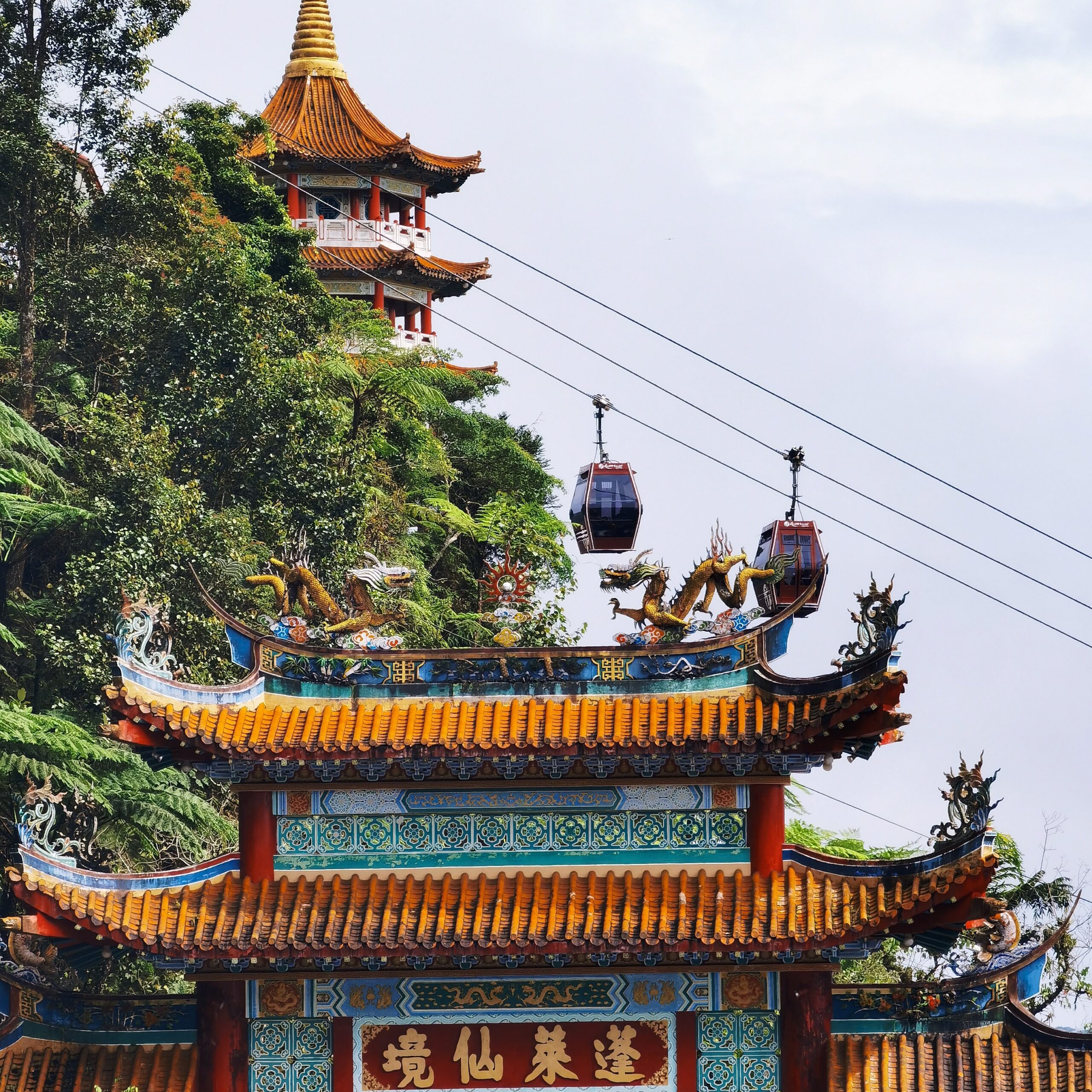 An ornate Chinese temple gateway with golden dragons, with a hillside pagoda and modern cable cars in the background in Genting Highlands, Malaysia