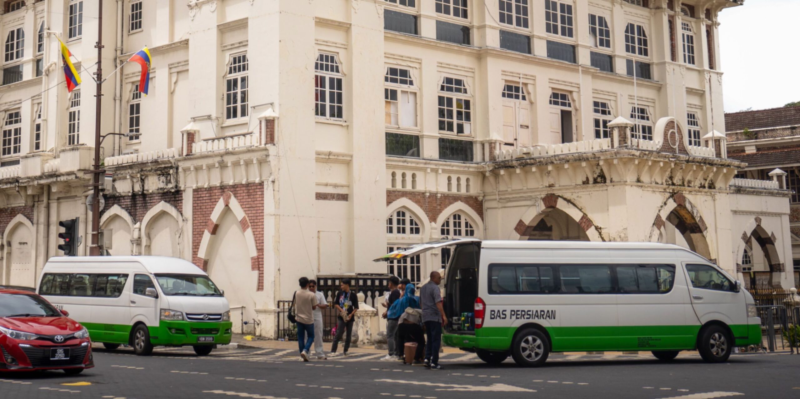 An old historic building with small tourist buses outside, small white and green vans.