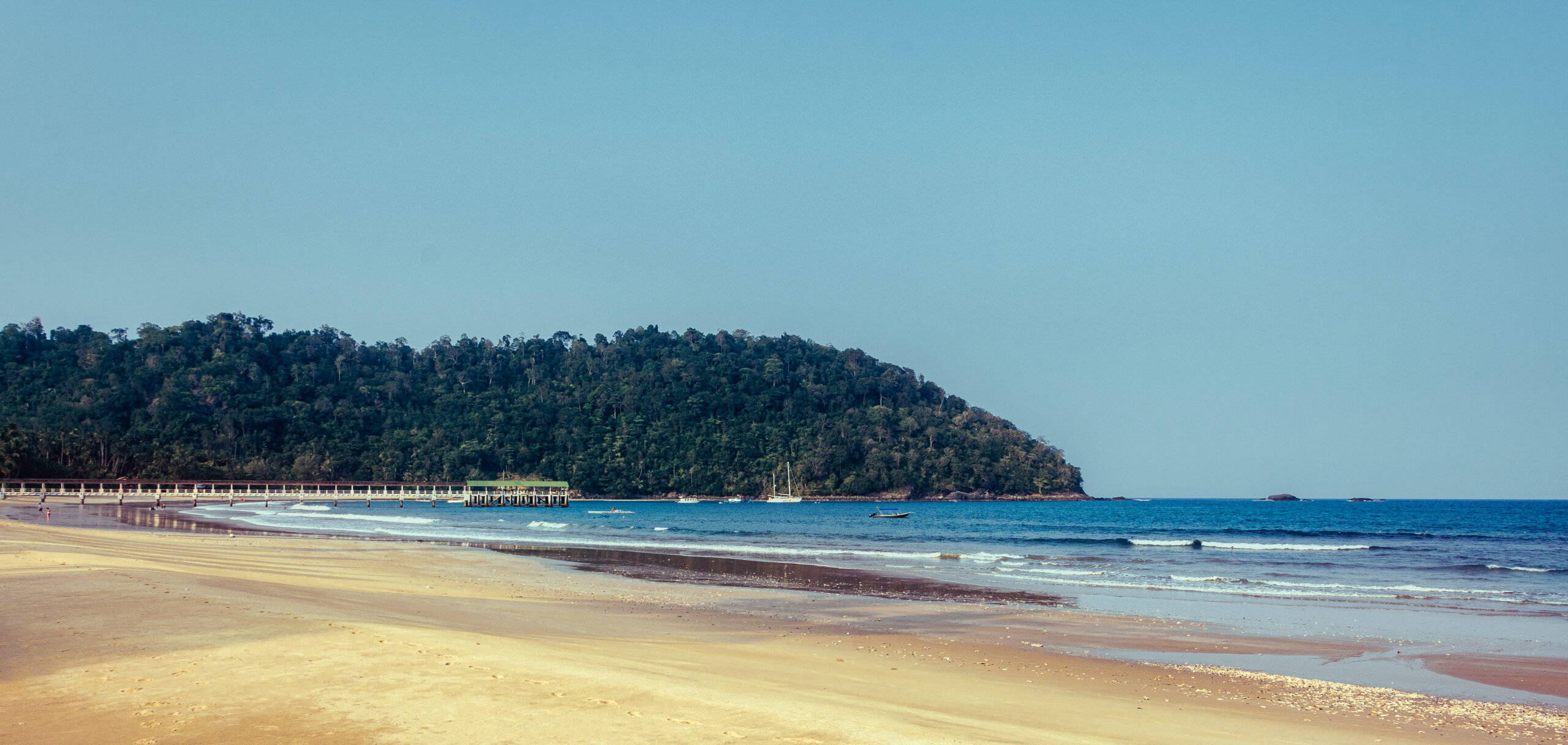 A wide sandy beach looking towards a dense, jungle-covered hill and a long coastal pier over a calm ocean.