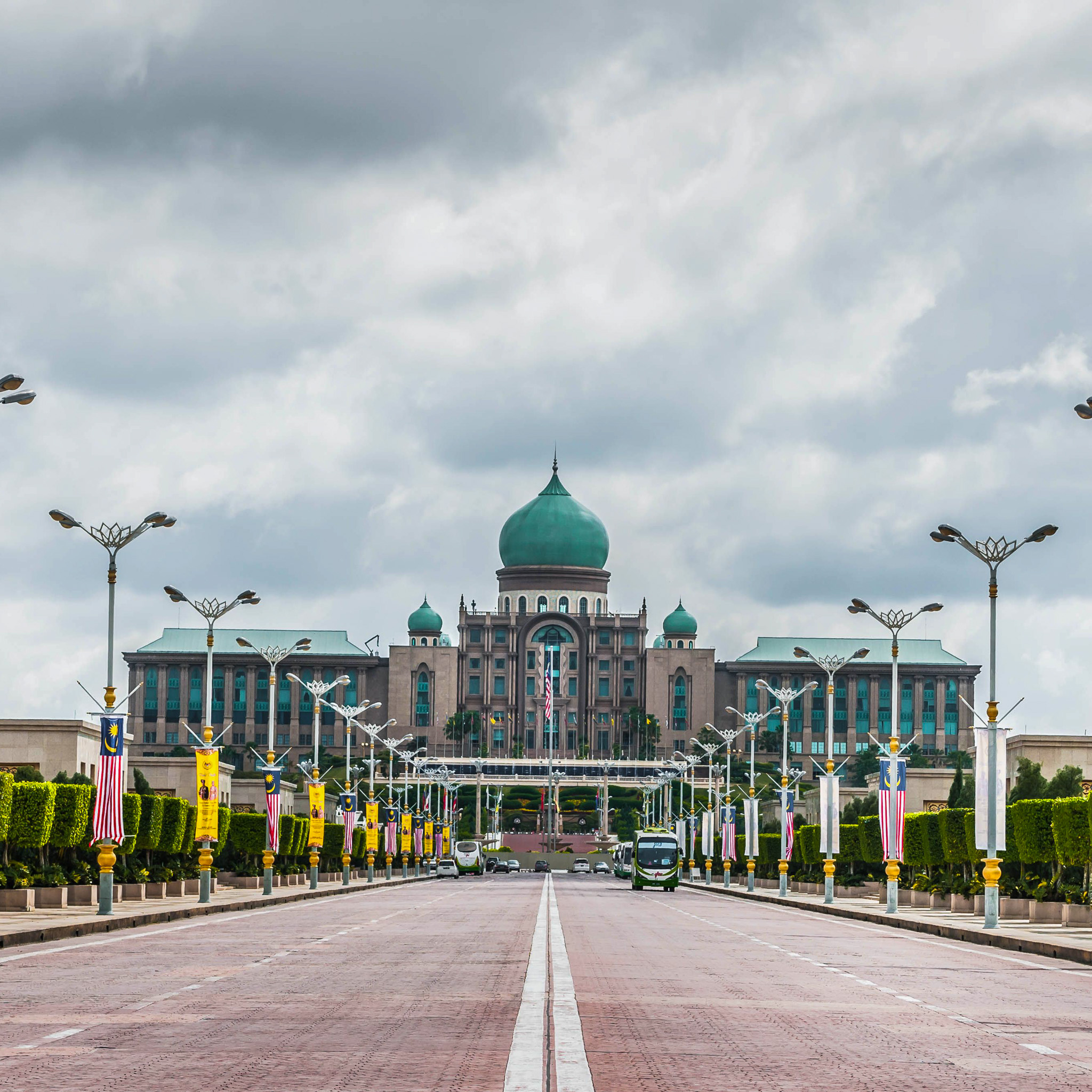 A wide, straight paved boulevard leads directly toward the grand, symmetrical facade of the Perdana Putra complex in Putrajaya, Malaysia. The massive stone building features a prominent central green onion dome alongside smaller flanking domes. The long road is lined on both sides by ornate, multi-pronged streetlights adorned with vertical Malaysian flags and yellow banners. Neatly trimmed green hedges border the walkways. The sky overhead is overcast with heavy, grey clouds, and a couple of buses are visible in the distance on the road.