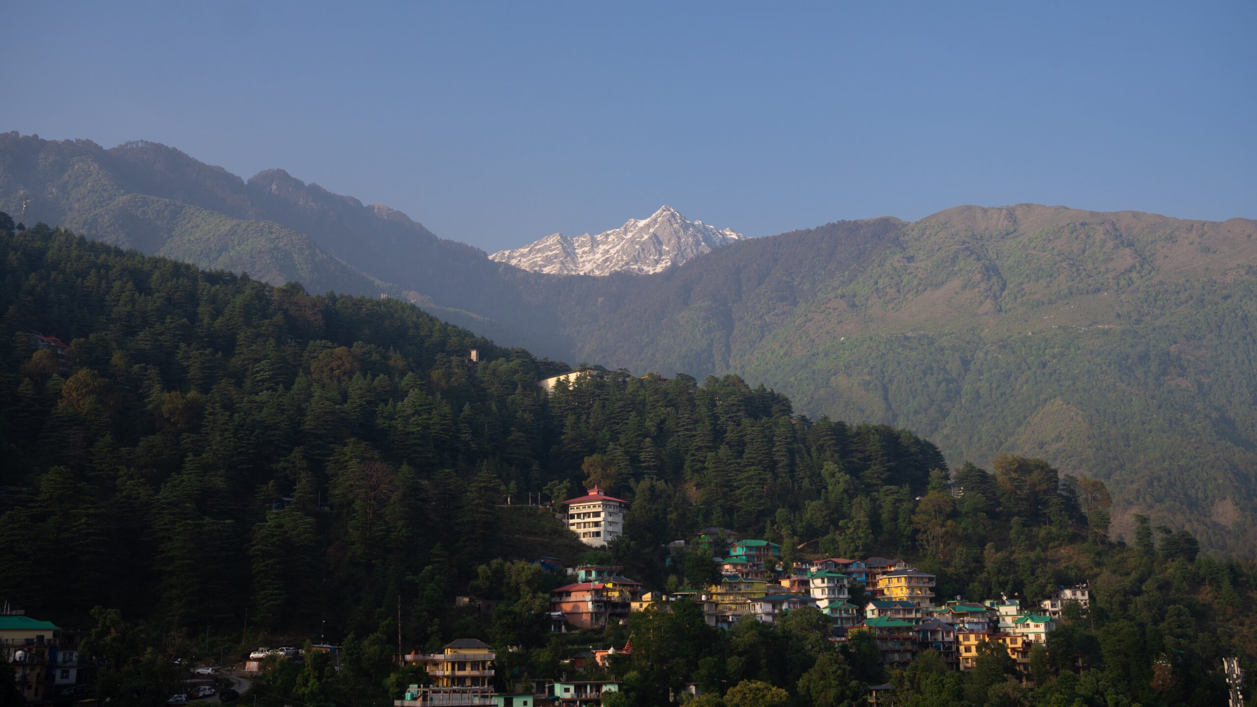 A snow-capped mountain in the middle of green hills and mountain houses below with a bright blue sky