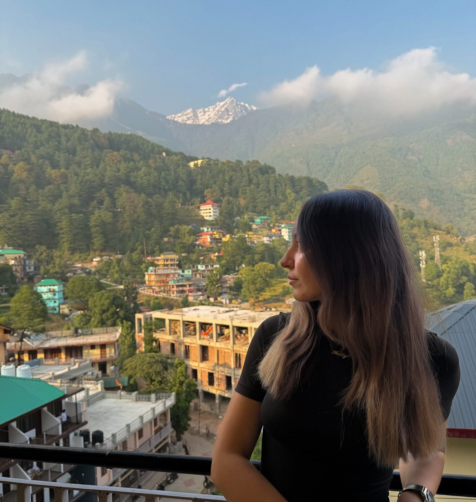 A women stood on a balcony with a mountain town in the background and mountains under the clouds in the distance
