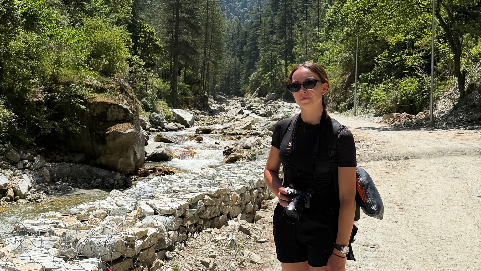 A women stood holding a camera, wearing hiking clothes on a hiking train with lush greenery and a bright sun.