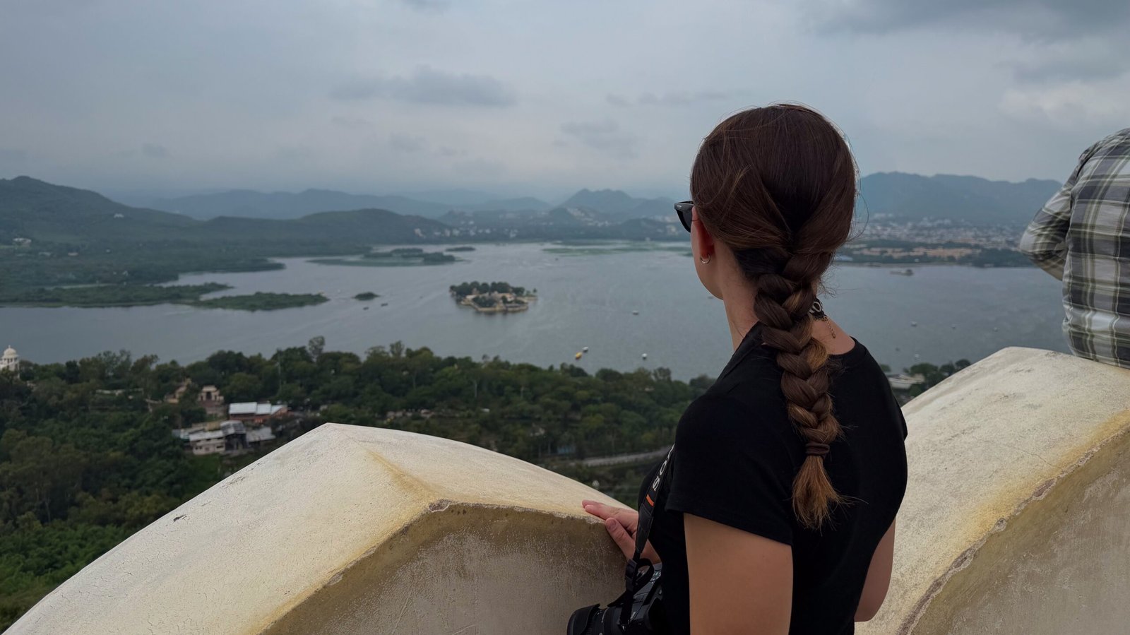 A women looking out over a wall to a view of a large lake with green hills in the background of Udaipur in India.