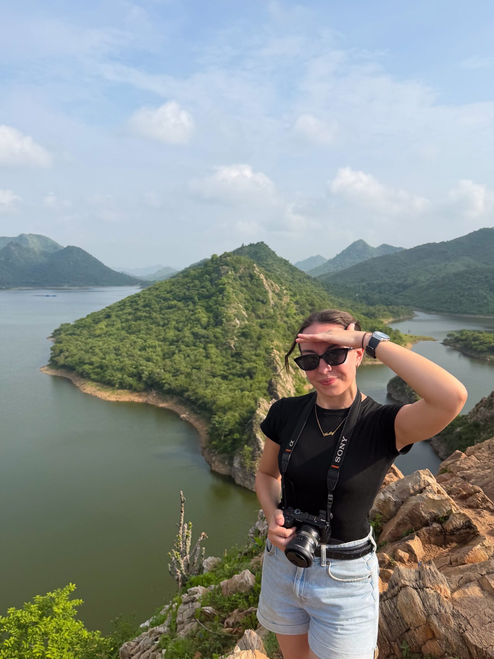 A women stood at a high elevation with a large green hill behind her, surrounded by a lake